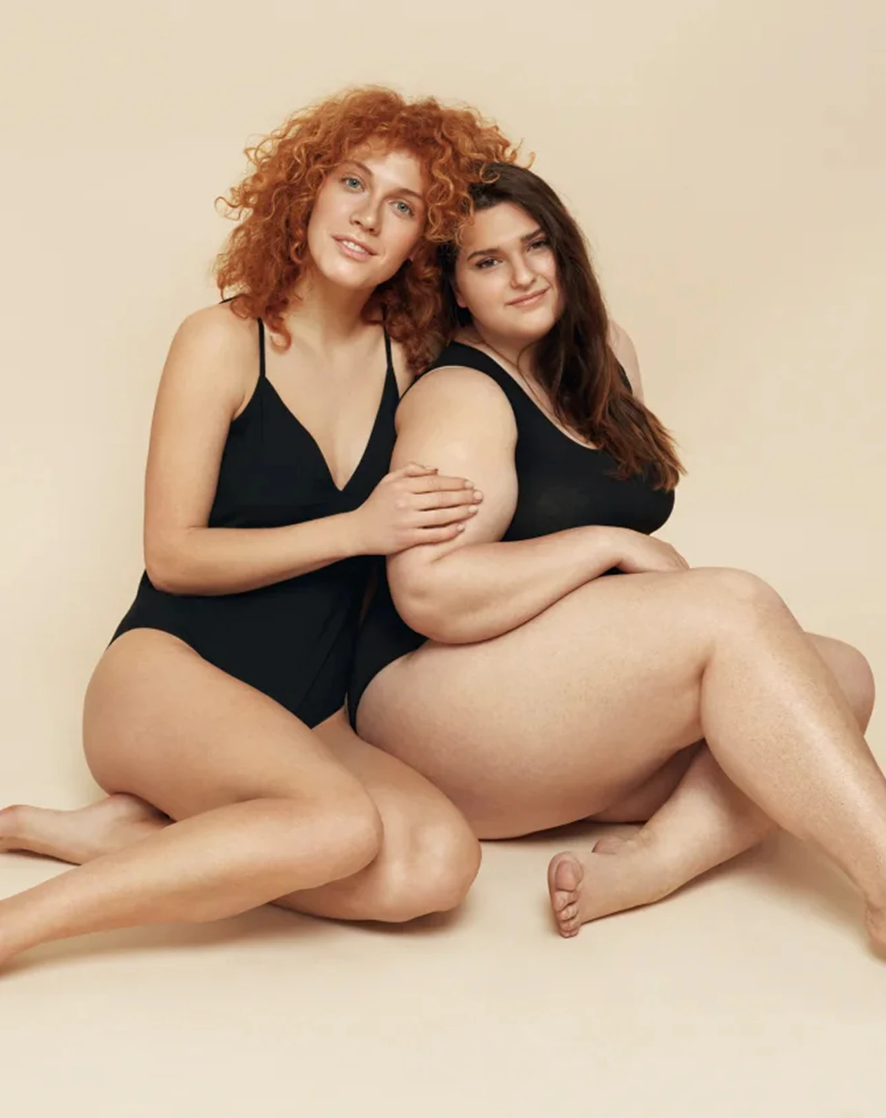 Dos mujeres en bañador, sentadas y abrazadas sobre fondo beige