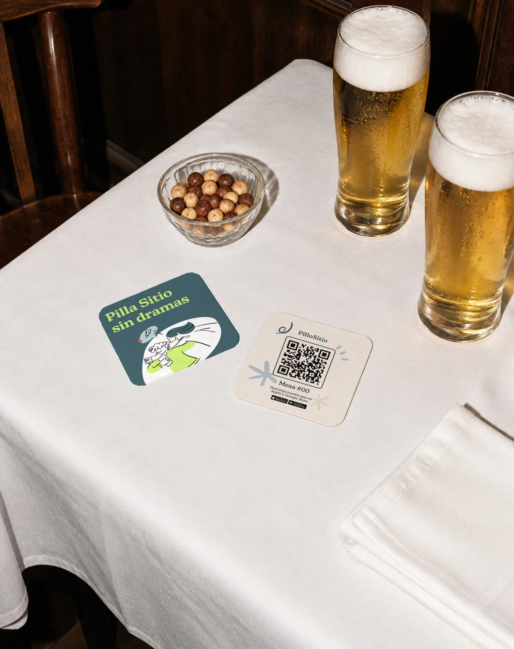 Posavasos de PilloSitio en la mesa de un bar, junto a dos cañas: «Pilla Sitio sin dramas» y el QR de la mesa