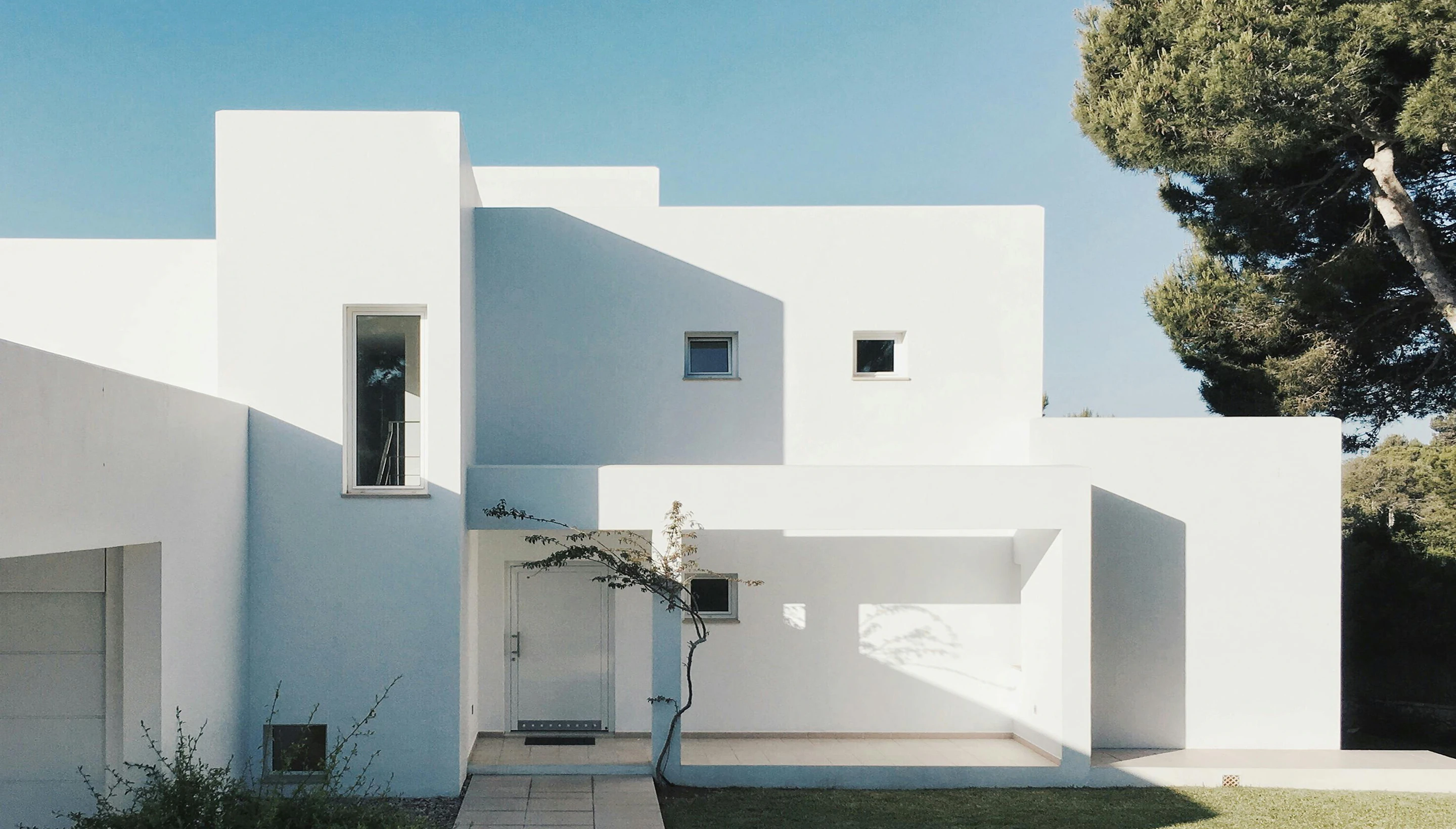 Una casa blanca de líneas rectas junto a un pino, bajo el cielo azul